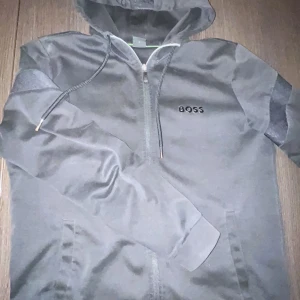 Äkta svart huvtröja från Boss  - Snygg svart huvtröja från Boss med dragkedja och justerbar huva. Tröjan har långa ärmar och ett diskret märke på bröstet. Ärmarna har en cool detalj med Boss-logotypen i ett mönster. Perfekt för en stilren look.