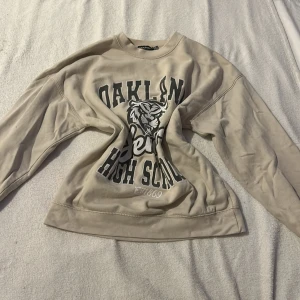 Beige sweatshirt från Boohoo - Säljer en beige sweatshirt från Boohoo med tryck 'Oakland Senior High School' och en tiger på framsidan. Tröjan har långa ärmar och en avslappnad passform. Perfekt för en casual look.