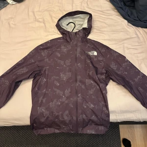 Lila regnjacka från The North Face stl S - Säljer en lila regnjacka från The North Face med ett snyggt mönster. Jackan har en dragkedja framtill och är långärmad med huva. Perfekt för regniga dagar och har en praktisk ficka på baksidan.