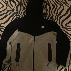 Grå och svart huvtröja från Nike - Säljer en snygg grå och svart huvtröja från Nike. Tröjan har en dragkedja framtill och två praktiska fickor med dragkedjor. Den har en klassisk Nike-logga på bröstet och är perfekt för en sportig look. Nästan helt oanvänd 