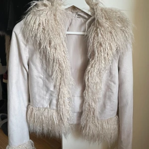 Beige pälsjacka med fuskpälsdetaljer - Säljer en snygg beige jacka med fluffiga fuskpälsdetaljer på krage och nederkant. Jackan har en stilren design med långa ärmar och en mjuk känsla. Perfekt för att hålla sig varm och trendig under kyliga dagar.
