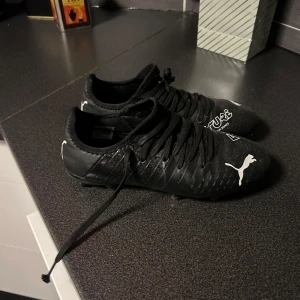 Svarta fotbollsskor från Puma - Snygga svarta fotbollsskor från Puma med vit logga och texten 'FUTURE' på ovansidan. Skorna har snörning och dobbar för bra grepp på planen. Perfekta för fotbollsträning och matcher. 