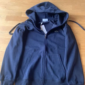 Mörkblå hoodie från Lacoste - Säljer en snygg mörkblå hoodie från Lacoste med dragkedja och den klassiska krokodilloggan på bröstet. Perfekt för en avslappnad stil. Pris kan diskuteras vid snabb affär