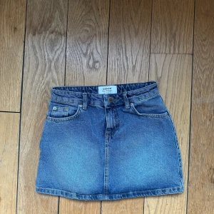 Blå jeanskjol - Snygg blå jeanskjol med klassisk femficksdesign. Kjolen har en kort längd och en knapp- och dragkedjestängning framtill. Säljer pga för liten för mig💙💙💙