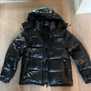 Svart pufferjacka från Polo Ralph Lauren - Säljer en snygg svart pufferjacka från Polo Ralph Lauren med en röd logga på bröstet. Jackan har en praktisk huva och dragkedja framtill. Perfekt för kyliga dagar! Storlek M. Endast använd en gång då den var för stor för mig!