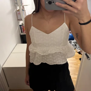Vit ärmlös topp med volang - Säljer en söt vit ärmlös topp med smala axelband och volangdetaljer. Toppen har ett diskret blommönster och är perfekt för sommardagar. Den är luftig och bekväm att bära.