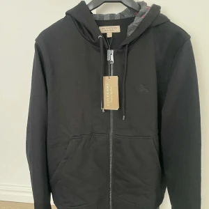 Svart hoodie från Burberry - Säljer en stilren svart hoodie från Burberry med dragkedja och justerbar huva. Insidan har ett klassiskt Burberry-rutigt mönster i grått och rött. Perfekt för en casual look. 