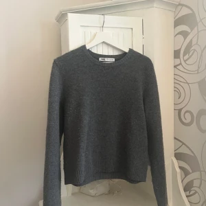 Grå stickad tröja från Zara - Säljer en stilren grå stickad tröja från Zara. Tröjan har en klassisk rund hals och långa ärmar, perfekt för kyligare dagar. Den är mjuk och bekväm med ribbade kanter vid ärmslut och nederkant.