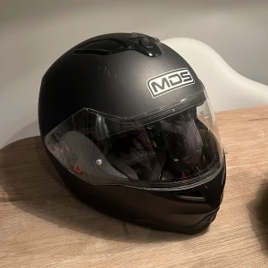 Svart hjälm från MDS - Säljer en svart hjälm från MDS med visir. Hjälmen har en stilren design, dock små märken på sig. . Den har ventilationsöppningar för bättre luftflöde och komfort samt inbyggda ”solglasögon” så man inte blir bländad. Man kan även öppna upp hjälmen som ni ser på sista bilden. Änvänd under 1 sommar. 