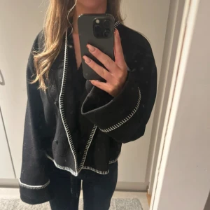 Svart kappa med vita detaljer - Snygg svart kappa med kontrasterande vita sömmar. Den har en oversized passform och långa ärmar. Perfekt för att ge en stilren look till din outfit.( lånad bild) storlek S(36)