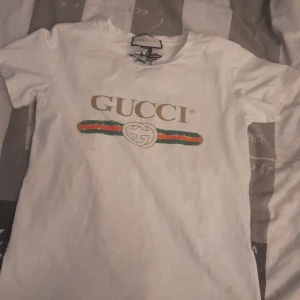 Vit t-shirt från Gucci - Säljer en vit t-shirt från Gucci med det klassiska logotyptrycket i guld och grönt/rött band på framsidan. T-shirten är kortärmad och tillverkad i bomull. Perfekt för en stilren look.