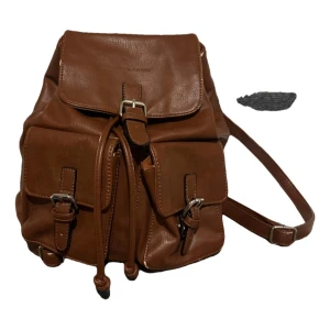 David Jones ryggsäck i skinn-imitation  - Snygg brun ryggsäck i skinn-imitation (polyuretan)  från David Jones. Den har två framfickor med spännen och en praktisk dragsko för att hålla dina saker säkra. Insidan är rymlig med en extra ficka med dragkedja för småsaker. Perfekt för en stilren look! Använd skick med lite skavanker i kanterna som ger en charmig vintage känsla. 