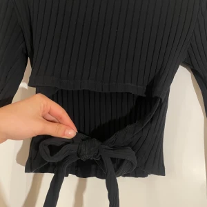 Svart ribbad topp med knyt - Snygg svart ribbad topp med långa ärmar och knytning i midjan. Perfekt för en stilren look.