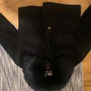 Svart kappa med pälsdetalj - Snygg svart kappa med en lyxig pälsdetalj runt kragen. Jackan har stora fickor framtill och knäpps med knappar. Perfekt för kyligare dagar när du vill hålla stilen. Köpte på Chelsea 