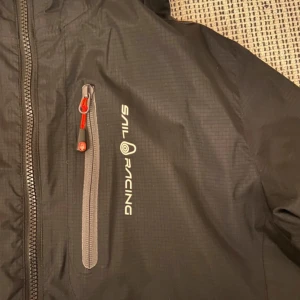 Svart jacka från Sail Racing - Säljer en svart jacka från Sail Racing i storlek medium. Jackan är tillverkad av polyamid och polyester, perfekt för segling och andra utomhusaktiviteter. Den har en cool USA 77-patch och dragkedja framtill. Perfekt för höst och vår!