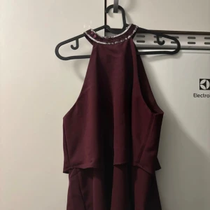 Vinröd klänning med paljettdetaljer - Snygg vinröd topp  i ett mjukt material med glittriga paljetter runt halsen. Den har en elegant knytning i nacken och är ärmlös, perfekt för festliga tillfällen. Toppen har en lös passform som ger en bekväm känsla.