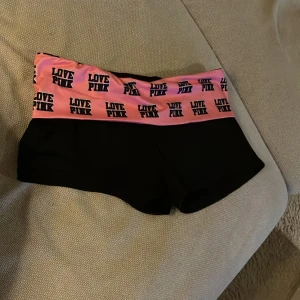 Svarta shorts med rosa resår  - Säljer ett par svarta shorts med en bred rosa resår i midjan där det står 'LOVE PINK' i svart text. Perfekta för en avslappnad dag eller träning. De är i bra skick och superbekväma att ha på sig. Passar perfekt för sommarens alla aktiviteter!