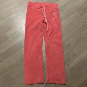 Rosa mjukisbyxor från Juicy Couture - Säljer ett par supermysiga rosa mjukisbyxor från Juicy Couture. De har en snygg broderad logga med glittriga detaljer på ena benet och en bekväm dragsko i midjan. Perfekta för en chill dag hemma eller en avslappnad promenad. 🩷