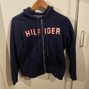 Mörkblå hoodie från Hilfiger - Säljer en snygg mörkblå hoodie från Hilfiger med dragkedja och stor logga på bröstet. Den har en skön passform och är perfekt för både vardag och chill. Fickor fram och justerbar huva gör den extra praktisk. Perfekt för höst och vår!
