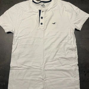 Vit t-shirt från Hollister - Säljer en vit t-shirt från Hollister i bra skick. Den har korta ärmar och en knappdetalj vid halsen. Perfekt för sommardagar eller avslappnade tillfällen. Klassisk och stilren design med Hollisters logga på bröstet. Nypris 200:-