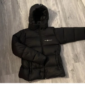 Svart dunjacka från Sail Racing  - Säljer en svart dunjacka från Sail Racing i relativt bra skick. Lite lagningar på vissa stället. Den är perfekt för vintern med sin varma och puffiga design. Jackan har en dragkedja framtill och en mysig huva för extra skydd mot kylan. Perfekt för kalla dagar! 🖤 Pris kan diskuteras!