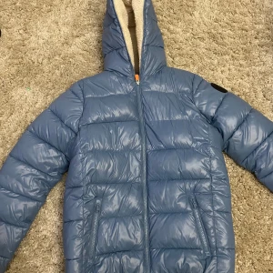 Blå vinterjacka save the duck - Säljer en snygg blå dunjacka från Save The Duck. Jackan har en mysig teddyfodrad insida och en praktisk huva. Perfekt för kalla vinterdagar! Den har en dragkedja framtill och två fickor med dragkedja. Jackan är i mycket bra skick och redo för nya äventyr. Den är nästan helt ny med väldigt bra skick, allt är perfekt.