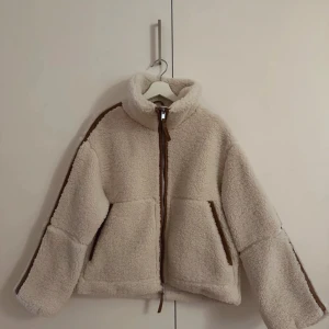 Beige teddyjacka med bruna detaljer - Mysig beige teddyjacka med bruna detaljer och dragkedja framtill. Perfekt för höst och vinter. Jackan har en hög krage och långa ärmar, vilket gör den supervarm och skön. 🤍🤎