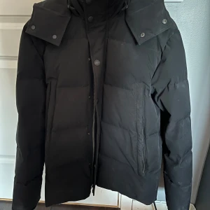 Svart pufferjacka med huva - Säljer en svart pufferjacka i mycket bra skick. Den har en praktisk huva och stängs med både dragkedja och knappar. Perfekt för kalla vinterdagar. Jackan är långärmad och har en stilren design som passar till det mesta. Perfekt för dig som vill hålla dig varm och snygg i vinter! Nypris ca 3000kr. Pris kan diskuteras så hör av dig vid minsta lilla funderingen!