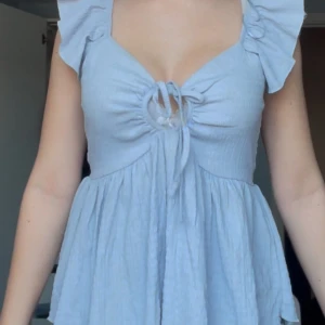 Ljusblå blus med volangdetaljer - Superfin ljusblå blus med volangdetaljer och knytning framtill. Perfekt för sommardagar eller en mysig vårutflykt. Den har en söt offshoulder-stil som ger en romantisk touch.