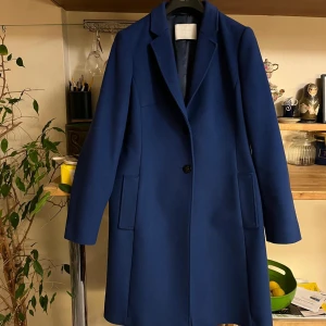 Blå kappa BOSS - Säljer en elegant blå kappa i ullblandning. Den har en klassisk design med en enkel knapp framtill och långa ärmar. Perfekt för höst och vinter, och ger en stilren look till vilken outfit som helst. Passar både till vardags och festliga tillfällen.