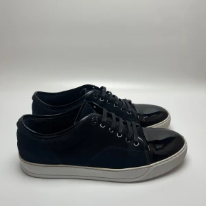 Lanvin skor  - Säljer ett par stilrena mörkblåa sneakers från Lanvin i bra skick. Skorna har en blank tå och snörning, vilket ger dem en elegant touch. Perfekta för både vardag och fest! Sulan är vit och har lite slitage, men inget som påverkar användningen. Passar perfekt till jeans eller en mer uppklädd stil. Uk 10 passar 45