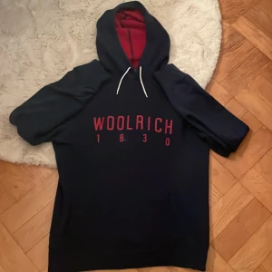 Mörkblå hoodie från Woolrich - Säljer en snygg mörkblå hoodie från Woolrich med röd text på framsidan. Den har en  ficka fram och en  huva med vita snören. Perfekt för kyliga dagar och passar till det mesta. Använd ett fåtal gånger, så den är i bra skick. 