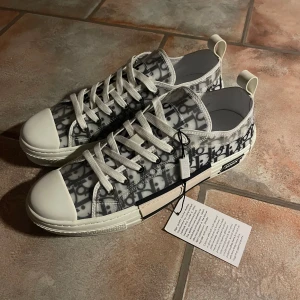Dior sneakers med monogrammönster - Säljer ett par snygga Dior sneakers i nytt skick. De har ett coolt monogrammönster i grått och vitt med vita snören och tåhätta. Skorna kommer i originalförpackning och är perfekta för att ge din outfit en lyxig touch. Passar både till vardags och fest!