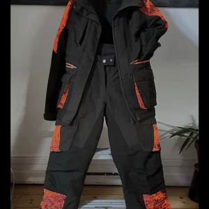 Svart och orange jacka med byxor (Pinewood) Str byxor 36 Jacka 38 - Pinewood super skick. Jakt/fiske ställ. Säljer en cool svart och orange jacka med matchande byxor. Perfekt för utomhusaktiviteter med sitt slitstarka material och praktiska fickor. Jackan har en dragkedja och huva, och byxorna har flera fickor för förvaring. Det orange mönstret ger en snygg kontrast mot det svarta.