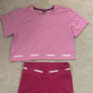 Rosa träningsset från Stronger🌸🏋🏼‍♀️ (priset inkluderar båda plaggen) - Säljer ett snyggt rosa träningsset från Stronger. Toppen är croppad med korta ärmar och har texten 'STRONGER' längs fållen. Byxorna är i matchande rosa med vita detaljer på benet och texten 'STRONGER' i midjan. Perfekt för gymmet eller yoga!