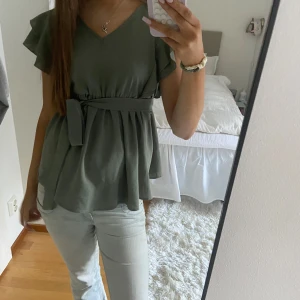 Grön blus  - Säljer en söt grön blus med volangärmar och knytband i midjan. Blusen har en v-ringad hals och är perfekt för både vardag och fest. Den är i mycket bra skick och passar bra till både jeans och kjol.