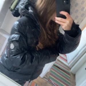 Svart pufferjacka från Moncler - Säljer en snygg och varm svart pufferjacka från Moncler. Jackan har en glansig finish och är perfekt för kalla vinterdagar. Den har en huva och långa ärmar, vilket gör den supermysig. Den har en liten defekt på framsidan av jackan❤️skriv för fler bilder