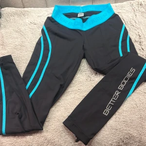 Svarta träningsleggings från Better Bodies - Säljer ett par svarta träningsleggings från Better Bodies med turkosa detaljer och logga längs benet. De är i bra skick och perfekta för gymmet eller löprundan. Midjan är elastisk och bekväm, vilket ger en bra passform under träningen. Dom är i ny skick och är i storleken XS för kvinnor