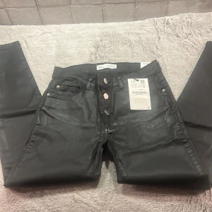 Svarta skinny jeans från Zara - Säljer ett par svarta skinny jeans från Zara i storlek 152. De är i nytt skick med etikett kvar. Jeansen har en snygg glansig finish och knappar i silverfärg. Perfekta för både vardag och fest!