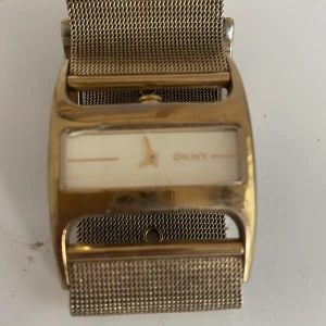 Guldfärgad klocka från DKNY - Säljer en stilren guldfärgad klocka från DKNY med en elegant mesh-armband. Klockan har en rektangulär urtavla i vitt och guld, vilket ger den en modern och minimalistisk look , lite färg bortfall.Perfekt för både vardag och fest! ✨