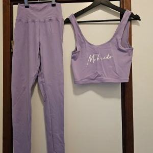Lila träningstopp och leggings från Makeido - Snyggt lila set med en croppad träningstopp och matchande leggings från Mkeido. Toppen har breda axelband och ett stilrent tryck framtill. Leggingsen är höga i midjan med rynkdetalj bak för en smickrande passform. Perfekt för yoga eller gymmet!
