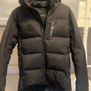 Svart dunjacka från Sail Racing - Säljer en svart dunjacka från Sail Racing i storlek medium. Jackan har en stilren design med dragkedja och logotypdetaljer på bröstet och ärmen. Perfekt för kalla vinterdagar med sin varma och bekväma passform. Huvan är justerbar och ger extra skydd mot väder och vind. Inköpt i november 2023, endast använd nov- mars. 