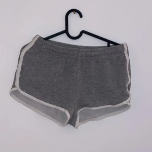Grå shorts med vita detaljer - Säljer ett par grå shorts med vita kanter. De är superbekväma och perfekta för en avslappnad dag hemma eller en snabb träningspass. Shortsen har en elastisk midja för extra komfort. De är i bra skick men har en liten slitage på kanten. Perfekta för sommaren!
