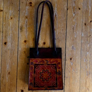 Vintage axelväska med blommönster - Säljer en unik vintage axelväska i skinnmaterial med ett vackert blommönster i rött och orange. Väskan har långa handtag och en praktisk dragkedja upptill. Har en ficka på utsidan med knapp, och en ficka på insidan med dragkedja. Perfekt för höst och vår, och ger en bohemisk touch till din outfit. Passar både till vardags och speciella tillfällen. Köparen står för frakt, skickar spårtbart PostNord paket.