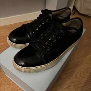 Svarta sneakers från Lanvin - Säljer ett par snygga svarta sneakers från Lanvin i mycket bra skick. Skorna har en stilren design med glansig finish och snörning. Perfekta för både vardag och fest! De har en bekväm passform och en vit sula som ger en cool kontrast. Kommer i originalkartong.har även kvitto