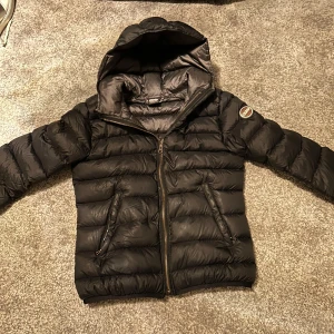 Svart dunjacka från Colmar - Säljer en snygg svart dunjacka från Colmar i mycket bra skick. Jackan har en huva och dragkedja framtill, perfekt för kyliga dagar. Den är quiltad och har en stilren design som passar både till vardags och lite finare tillfällen. Superbekväm och håller dig varm hela vintern!