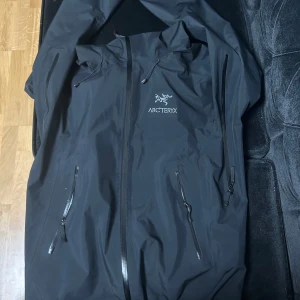 Svart vindjacka från Arc'teryx - Säljer en svart vindjacka från Arc'teryx i storlek S. Jackan har en stilren design med dragkedja och justerbar huva. Perfekt för blåsiga dagar och utomhusaktiviteter. Den är i mycket bra skick och redo för nya äventyr!