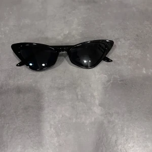 Svarta solglasögon i cat-eye stil - Snygga svarta solglasögon med en cool cat-eye form. Perfekta för att ge din outfit en edgy touch. De har en glansig finish och mörka linser som skyddar mot solen. Passar både till vardags och fest!