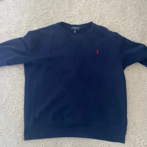 Mörkblå tröja från Polo Ralph Lauren - Säljer en snygg mörkblå tröja från Polo Ralph Lauren. Den har en klassisk design med ett rött broderat logotypmärke på bröstet. Tröjan är i storlek 14-16 år(XS) och är perfekt för både vardag och lite finare tillfällen. Superbekväm och stilren!