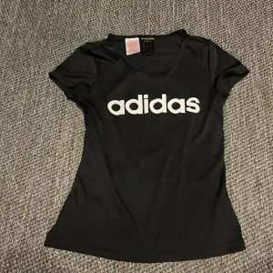 Säljer en svart t-shirt från Adidas med deras klassiska logga tryckt i vitt på framsidan. T-shirten är kortärmad och har en normal passform. Perfekt för träning eller vardagsbruk!